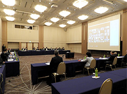 ウェブシステム併用会議のテスト兼ね開催された幹事会で、スクリーンに映し出されたオンライン参加者を見ながら挨拶する佐々木代表幹事（左側の列手前から３人目）